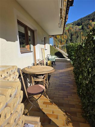 Een gezellige balkon met een ronde tafel en twee stoelen. Op de achtergrond zijn groene bergen en een heldere blauwe lucht te zien.