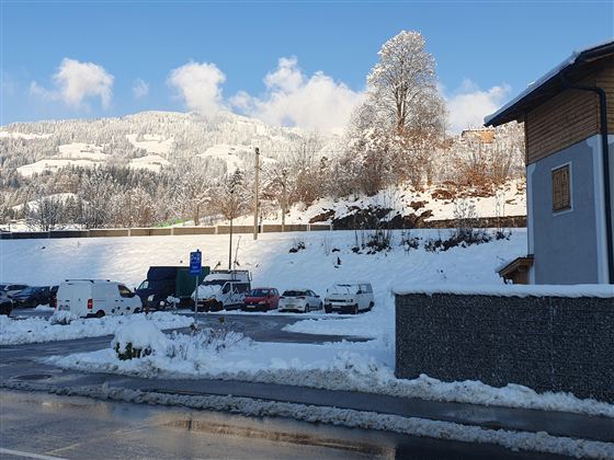 Eine verschneite Landschaft mit Parkplätzen und Bäumen im Hintergrund. Der Himmel ist klar und blau, was eine ruhige Winteratmosphäre schafft.