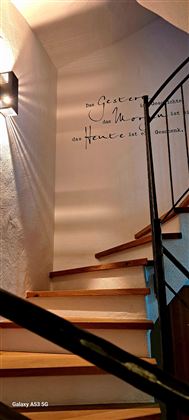 Eine elegante Treppe führt nach oben, beleuchtet von einer Wandleuchte. An der Wand ist ein inspirierendes Zitat geschrieben.