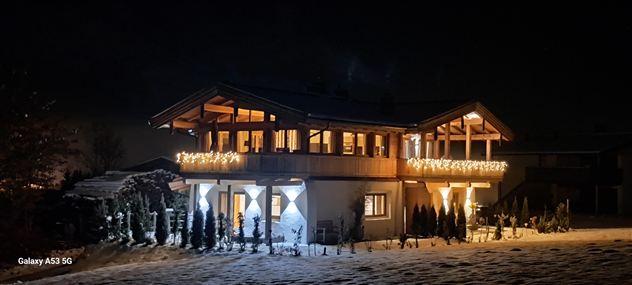 Ein gemütliches Haus in der Nacht, beleuchtet mit festlichen Lichtern. Der Schnee im Vordergrund sorgt für eine winterliche Atmosphäre.