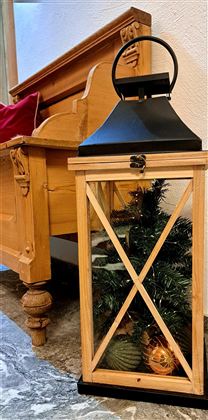 Eine Holzlaterne steht neben einer Holzbank und enthält eine kleine künstliche Tanne. Der Raum hat eine gemütliche Atmosphäre mit warmen Farben.