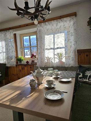 Ein gemütlicher Essbereich mit einem Holztisch, der liebevoll gedeckt ist. In den Fenstern sind helle Vorhänge und einige Pflanzen zu sehen.