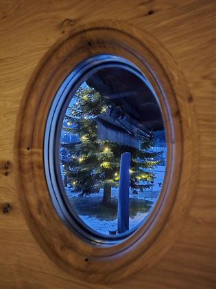 Ein runder Ausschnitt zeigt einen beleuchteten Weihnachtsbaum vor einem Holzhaus. Die Umgebung ist in sanftes Blau getaucht.