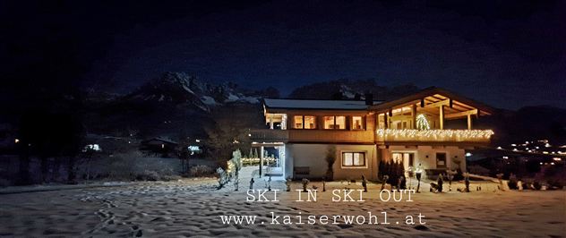 Ein gemütliches Chalet im Schnee bei Nacht. Im Hintergrund sind Berggipfel zu sehen, während die Lichter des Hauses strahlen.