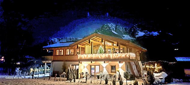 Ein schönes Holzhaus bei Nacht, umgeben von schneebedeckten Bergen. Das Gebäude ist festlich beleuchtet und strahlt eine gemütliche Atmosphäre aus.
