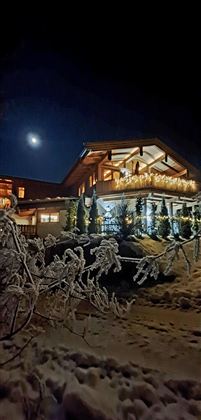 Ein winterliches Haus mit festlicher Beleuchtung unter einem klaren Nachthimmel. Der Schnee umgibt das Gebäude und der Mond scheint hell.
