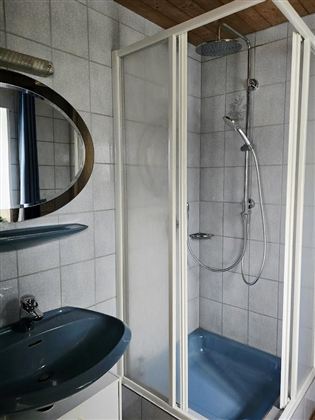 Ein modernes Badezimmer mit einer Duschkabine und einem Waschbecken. Die Fliesen sind hellgrau, und es gibt einen Spiegel über dem Waschbecken.