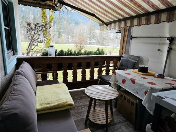 Ein gemütlicher Balkon mit einer Couch, einem kleinen Tisch und einer Tischdecke mit roten Punkten. Im Hintergrund sind Bäume und Berglandschaft sichtbar.