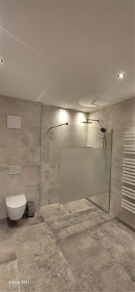 Ein modernes Badezimmer mit einer begehbaren Dusche und einem schlichten Design. Die Wände sind in hellen Farbtönen gehalten und der Boden besteht aus grauem Stein.