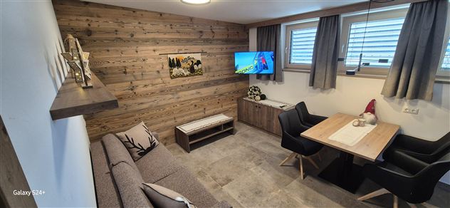 Ein gemütlicher Wohnraum mit Holzwänden und einem Fernseher. Ein Esstisch mit schwarzen Stühlen und einer Sofaecke lädt zum Entspannen ein.