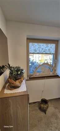 Ein gemütlicher Raum mit einem Fenster, durch das Sonnenlicht scheint. Auf einer Kommode steht eine Pflanze und ein kleiner Topf auf dem Boden.