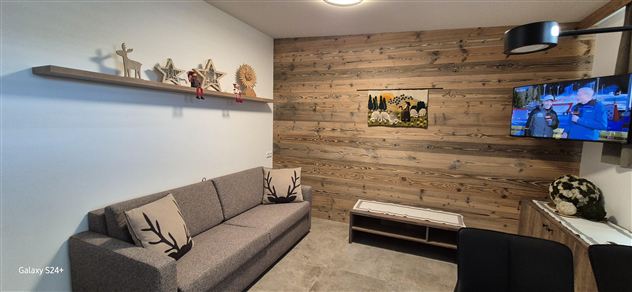 Ein gemütliches Wohnzimmer mit einem grauen Sofa und einer Holzwand. Ein Fernseher zeigt ein Programm und auf einem Regal stehen dekorative Elemente.