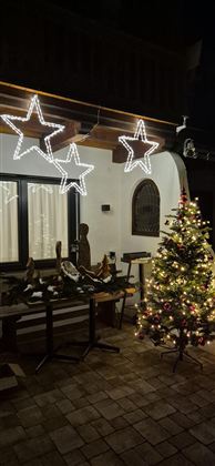 Eine festlich dekorierte Terrasse mit einem beleuchteten Weihnachtsbaum und leuchtenden Sternen. Im Hintergrund sind Fenster mit warmem Licht zu sehen.