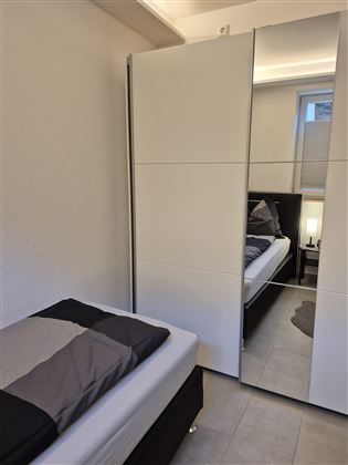 Ein modernes Schlafzimmer mit einem Einzelbett und einem großen Spiegelkleiderschrank. Die Wände sind hell und der Boden ist gefliest.