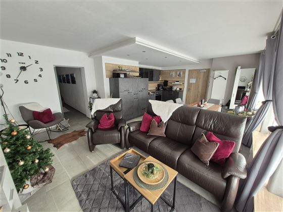 Ein modernes Wohnzimmer mit einem bequemen Sofa und zwei Sesseln. Im Hintergrund sind eine Küche und ein Weihnachtsbaum zu sehen.