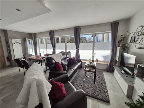 Ein modernes Wohnzimmer mit bequemen Möbeln und großen Fenstern. Der Raum ist hell und freundlich, mit dekorativen Elementen und einem gemütlichen Ambiente.