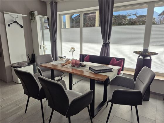 Ein moderner Essbereich mit einem Holz-Tisch und vier grauen Stühlen. Große Fenster lassen viel Licht herein und bieten einen Blick auf die Außenansicht.