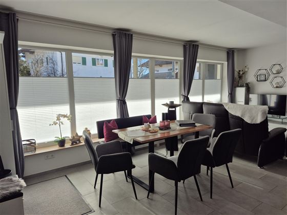 Ein modernes Esszimmer mit einem Holztisch und schwarzen Stühlen. Große Fenster mit Sonnenlicht, das den Raum erhellt.