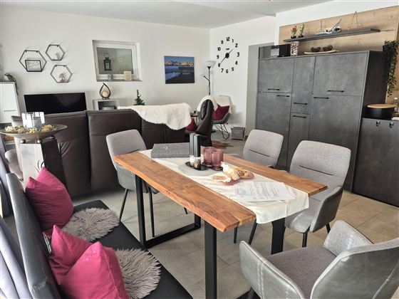 Ein modern eingerichteter Essbereich mit einem großen Holztisch und stilvollen Stühlen. Die Wände sind neutral, und es gibt dekorative Elemente wie Bilder und Pflanzen.