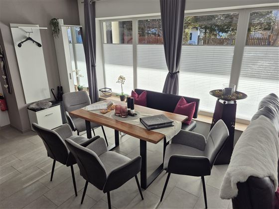 Ein modernes Esszimmer mit einem Holztisch und sechs Stühlen. Große Fenster lassen viel Licht herein und sorgen für eine helle Atmosphäre.