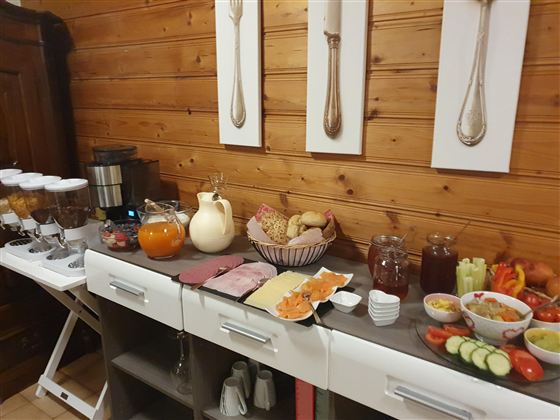 Ein reichhaltiges Frühstücksbuffet mit verschiedenen Aufschnittwaren, frischem Obst, Gemüse und Getränken. Der Hintergrund ist aus Holz, was eine gemütliche Atmosphäre schafft.