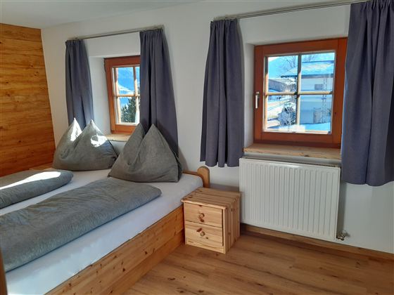 Pohodlná ložnice se dvěma postelemi a šedými polštáři. Okna propouští hodně denního světla a nabízejí výhled ven.