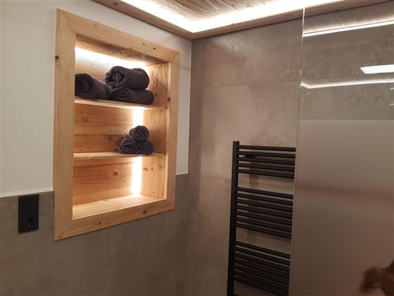 Moderní koupelna s dřevěnou policí, na které jsou uložené složené ručníky. Na zdi je viditelný radiátor na ručníky.