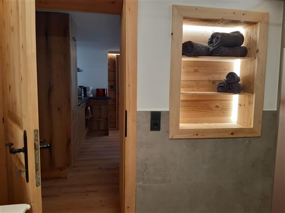 Moderní koupelna s dřevěnými policemi a složenými ručníky. V pozadí je viditelná otevřená kuchyně.