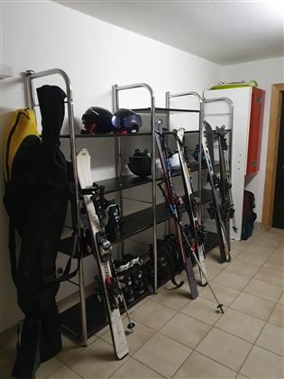 Ein Skiraum mit Regalen, auf denen Skiausrüstung, Helme und Skier aufgereiht sind. Die Wände sind neutral und der Boden ist gefliest.