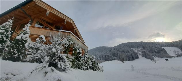 Ein gemütliches Holzhaus im verschneiten Winterlandschaft. Im Hintergrund sind bewaldete Hügel und ein bewölkter Himmel zu sehen.