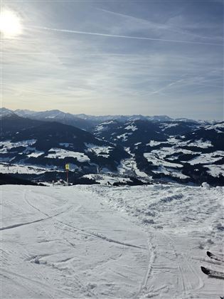 Eine winterliche Berglandschaft mit schneebedeckten Hügeln und mächtigen Bergen im Hintergrund. Der Himmel ist klar und die Sonne scheint.