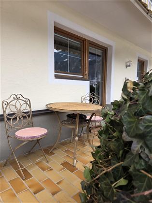 Ein kleine Terrasse mit einem runden Tisch und zwei Stühlen. Im Hintergrund sind eine Fenster und eine grüne Pflanze zu sehen.