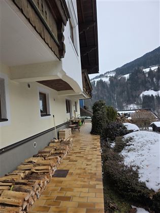 Ein schmaler Weg mit Holzstapel führt zu einem Gebäude in den Bergen. Schnee liegt auf dem Boden, und die Landschaft ist von Wäldern und Hügeln umgeben.