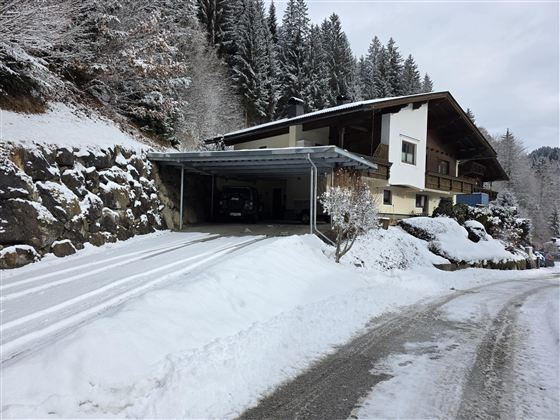 Een modern huis in de sneeuw, omringd door bomen. De oprit is bedekt met sneeuw en leidt naar de overdekte garage.