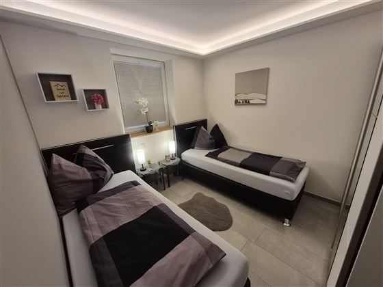 Ein modernes Schlafzimmer mit zwei Einzelbetten und einer eleganten Beleuchtung. Die Wände sind neutral gehalten und es gibt dekorative Elemente auf den Regalen.