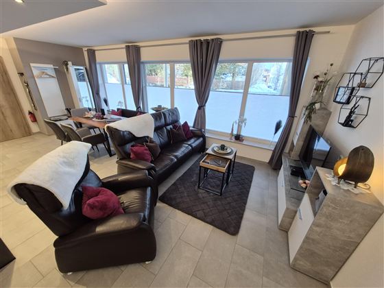 Ein moderner Wohnraum mit einem bequemen Sofa, einen Relaxsessel und großen Fenstern. Die Einrichtung ist warm und einladend, mit dekorativen Elementen.