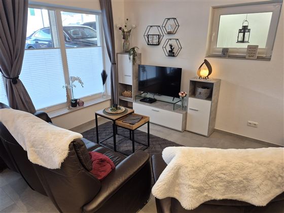 Ein gemütliches Wohnzimmer mit einem Ledersofa, einem Relaxsessel und einem Fernseher. Der Raum ist hell und modern eingerichtet, mit Pflanzen und dekorativen Elementen.