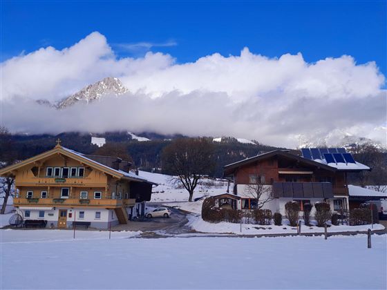 Een pittoresk winterlandschap met met sneeuw bedekte velden. Twee traditionele gebouwen staan voor majestueuze bergen en een bewolkte hemel.