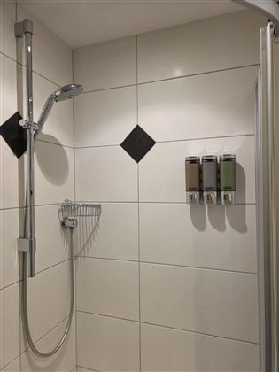 Een moderne douche met witte tegels en zwart decor. Aan de muur hangen dispensers voor shampoo en douchegel.
