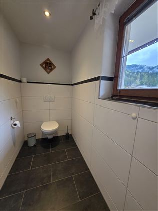 Een moderne kleine badkamer met witte tegels en een toilet. Door het raam is er een mooi uitzicht op de natuur zichtbaar.