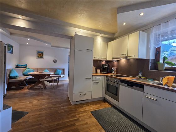 Eine moderne Küche mit hellen Schränken und Holzfußboden. Im Hintergrund ist ein gemütlicher Essbereich mit Sitzgelegenheiten zu sehen.