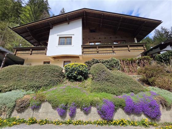 Ein zweigeschossiges Haus mit Holzverkleidung und Balkon. Der Garten ist mit bunten Blumen und Sträuchern bepflanzt.