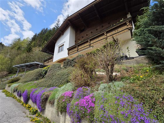 Ein hübsches Haus mit einem Balkon, umgeben von bunten Blumen. Im Hintergrund sind grüne Hügel und ein blauer Himmel zu sehen.