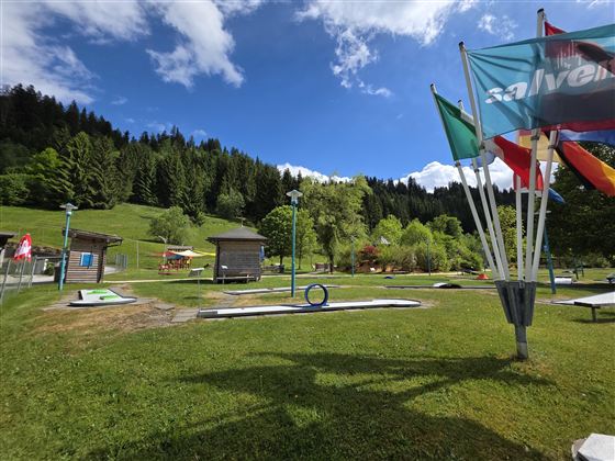 Een mooie minigolfbaan in de natuur met groene weiden en bomen op de achtergrond. Kleurrijke vlaggen wapperen in de wind onder een blauwe lucht.