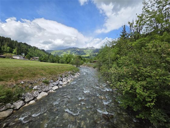 Een rustige rivier stroomt door een groene landschap met bergen op de achtergrond. De lucht is gedeeltelijk bewolkt en straalt een vredige sfeer uit.