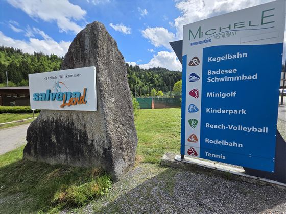 Een welkomstbord voor het vrijetijdsaanbod "salve4gol" naast een grote steen. Op de achtergrond staan vrijetijdsactiviteiten zoals midgetgolf, zwembad en beachvolleybal vermeld.