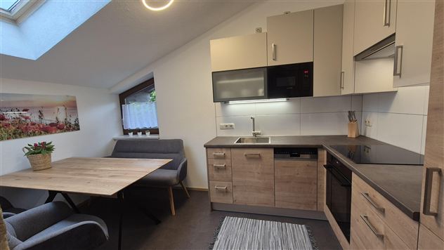 A modern kitchen with wooden shelves and light walls. The room has a large window and a small dining area with a table and chairs.