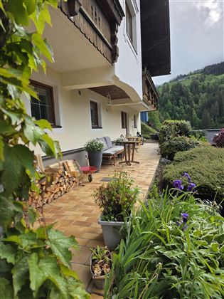 Een gezellige terras met planten en zitplaatsen. Op de achtergrond zijn groene heuvels en een heldere lucht te zien.