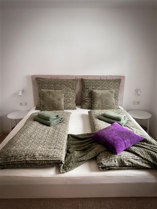 Een modern bed met twee kussens en verschillende dekens. Op het bed liggen groene handdoeken en een paarse kussen.