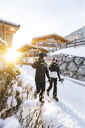 Zwei Skifahrer gehen einen verschneiten Weg entlang, umgeben von Chalets. Die Sonne scheint und sorgt für eine schöne Winteratmosphäre.